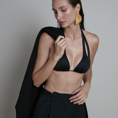 Mulher com top preto de alças finas, calças e blazer pretos, brincos amarelos grandes, fundo cinzento