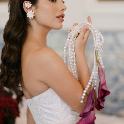 Mulher com vestido branco e brincos florais, segurando colar de pérolas com flores roxas