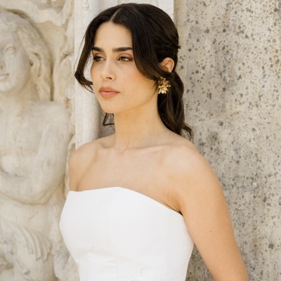 Mulher com vestido de noiva branco sem alças e brincos dourados com pérolas junto a parede de pedra.