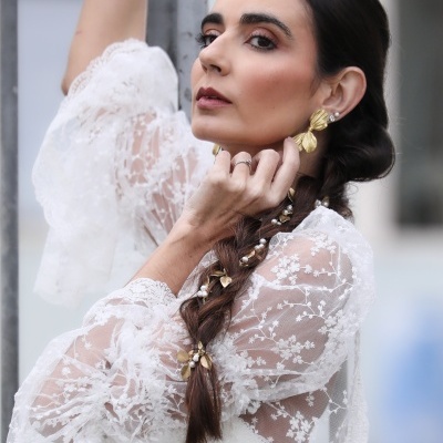 Mulher com blusa branca de renda translúcida e brincos dourados junto a grades metálicas cinza