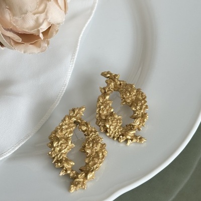 Brincos dourados texturizados ovalados sobre prato branco com flores creme ao lado