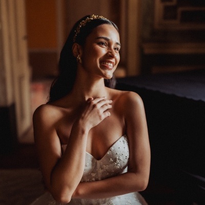 Mulher com vestido de noiva branco bordado sentada em ambiente interior com luz suave.