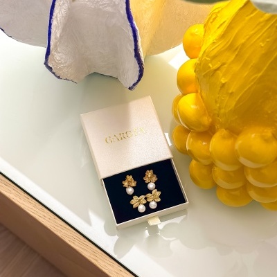 Brincos dourados com pérolas numa caixa branca GAROTA sobre mesa com vaso amarelo e decoração branca com borda azul.