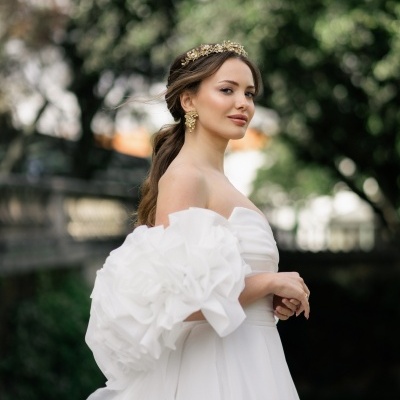 Mulher com vestido de noiva branco, flores de tecido num ombro, coroas douradas e brincos, em ambiente ao ar livre