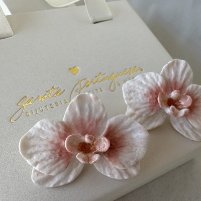 Brincos de orquídea rosa e branco sobre embalagem creme com texto dourado