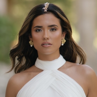 Mulher com vestido branco, brincos dourados com pérolas e presilha de cabelo decorativa.