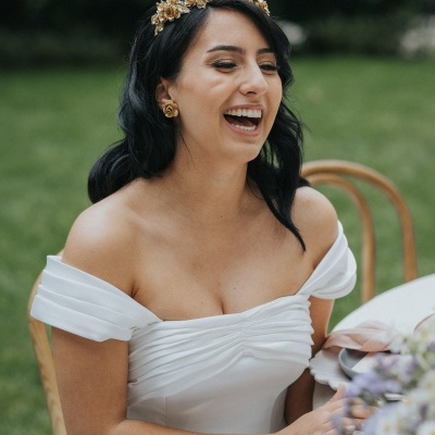 Mulher sorrindo com vestido de noiva branco e acessório dourado no cabelo, sentada em ambiente exterior