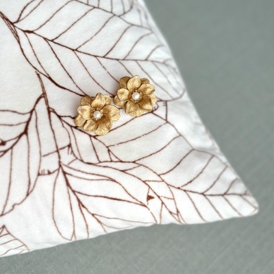 Brincos dourados em forma de flor com pérola sobre almofada branca com padrão castanho