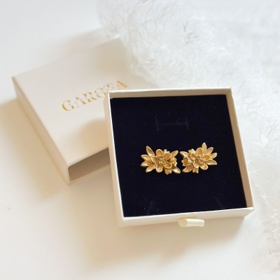 Brincos dourados em forma de flor numa caixa preta dentro de uma caixa branca com texto dourado GARÇON