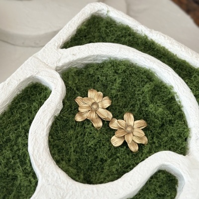 Brincos florais dourados sobre musgo verde e estrutura branca texturizada