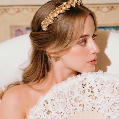 Mulher com tiara dourada com pérolas e brincos de flor dourados, segurando leque branco de renda com padrão floral.