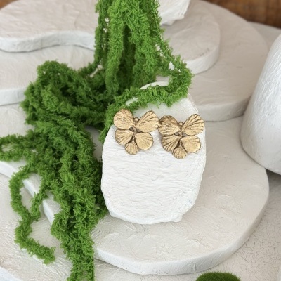 Brincos de flores douradas sobre pedestal branco com decoração verde ao redor.