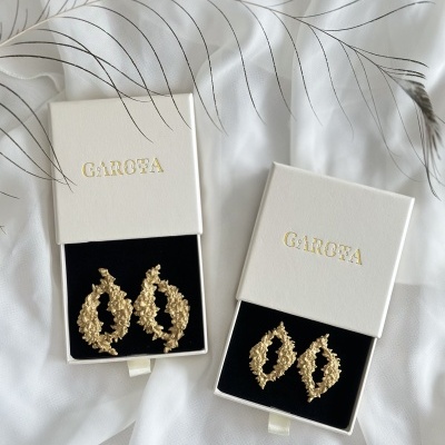 Brincos dourados em caixas brancas com texto GAROFA e pano branco com padrão de folhas cinza