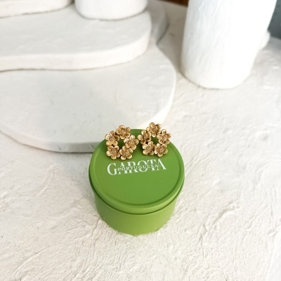 Brincos dourados de flores sobre caixa verde com texto branco em fundo branco texturizado