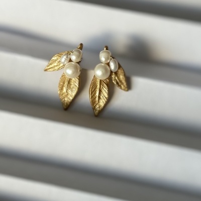 Brincos dourados com folhas e pérolas sobre fundo branco com sombras lineares