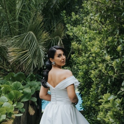 Vestido branco de noiva com decote ombro a ombro e botões nas costas em ambiente exterior com vegetação.