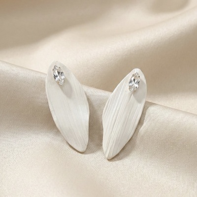 Brincos em forma de folha branca com pedra transparente sobre tecido bege.
