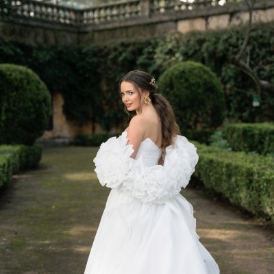 Mulher de vestido de noiva branco com mangas volumosas em jardim com arbustos e muro de pedra