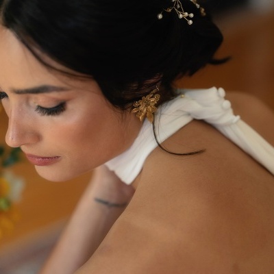 Mulher com vestido branco e brincos dourados florais com cabelo preso e flores ao fundo