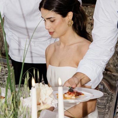 Mulher com vestidos branco e brincos dourados sentada à mesa com comida e velas