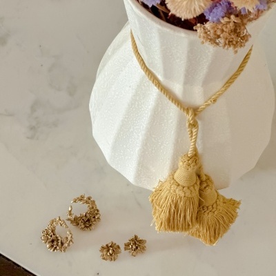 Vaso branco texturado com flores secas coloridas e cordão amarelo com franjas sobre superfície de mármore com brincos dourados ao lado.