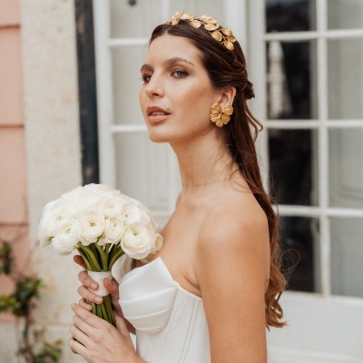 Mulher com vestido branco e acessórios dourados segurando bouquet de flores brancas