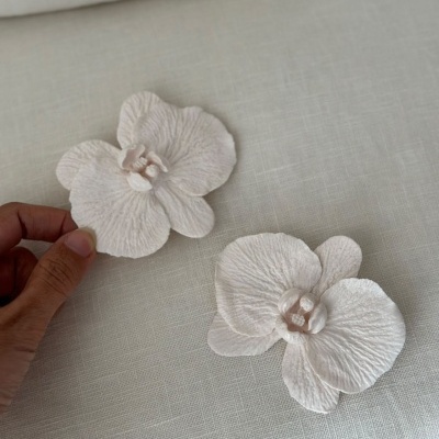 Dois acessórios florais brancos com textura sobre tecido claro, uma mão segura um deles