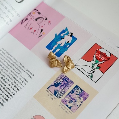 Brincos dourados com textura de folhas sobre revista aberta com ilustrações coloridas.
