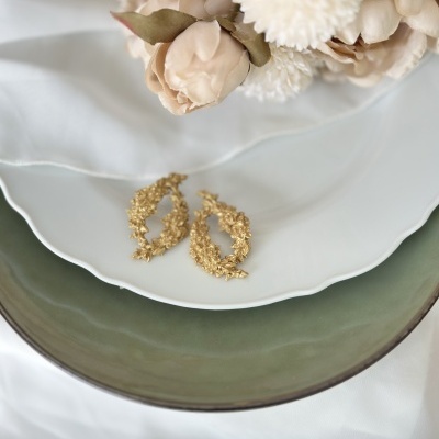 Brincos dourados sobre pratos branco e verde com flores artificiais ao fundo