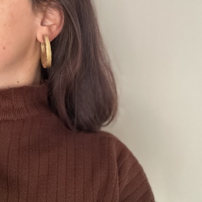 Brincos dourados de argola com textura, usados por pessoa com suéter castanho.