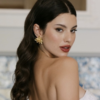 Brincos dourados em forma de flor usados por mulher com cabelo castanho ondulado e top branco.