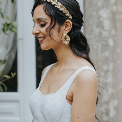 Mulher com vestido branco e acessórios dourados de flor, cabelo preso, em frente a porta de vidro