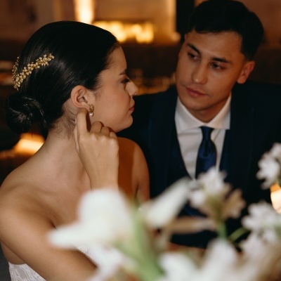 Mulher de vestido branco com acessórios dourados e homem de fato azul escuro num ambiente interior com flores brancas em primeiro plano