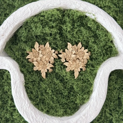 Brincos dourados com design floral sobre fundo verde texturado e moldura branca.