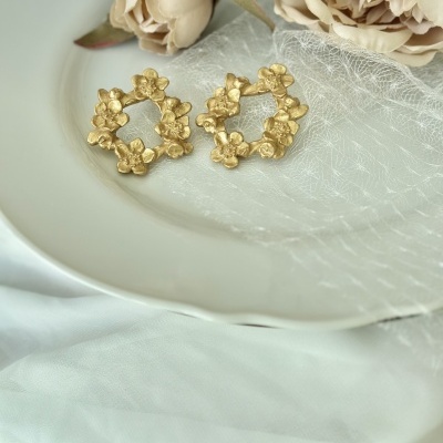Brincos dourados em forma circular com flores em relevo sobre prato branco com fundo de tecido e flores creme