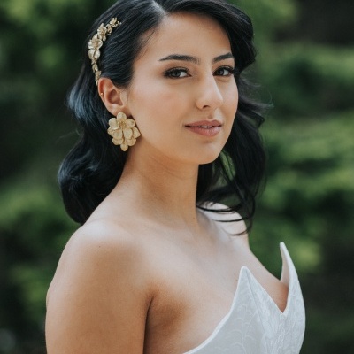 Mulher com vestido branco, cabelo preto com acessório dourado, brincos florais dourados, fundo verde desfocado