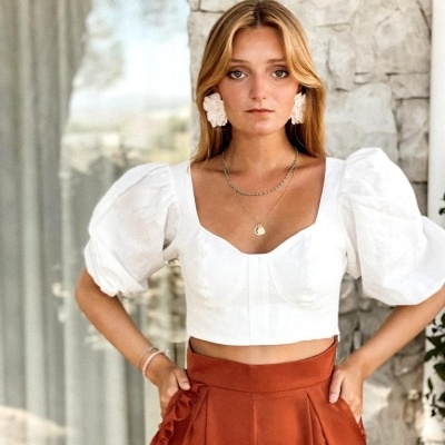 Modelo com blusa branca de mangas bufantes e calças ferrugem, em fundo de parede de pedra e vidro