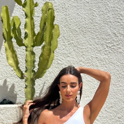 Mulher com blusa branca de um ombro e recorte, brincos grandes brancos, parede branca texturizada e cacto verde ao fundo.