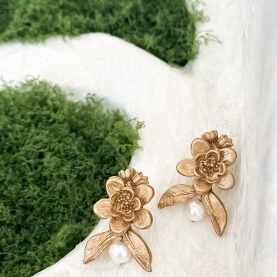 Brincos dourados com flores e pérolas sobre superfície branca e musgo verde