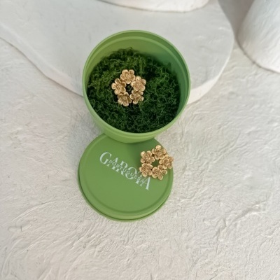 Brincos dourados em forma de flores dentro e fora de caixa verde redonda com texto GARDEN GARSON