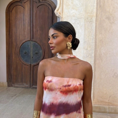 Mulher com vestido multicolor, brincos dourados em forma de folhas e braceletes dourados largos, em frente a porta de madeira e parede de mármore.
