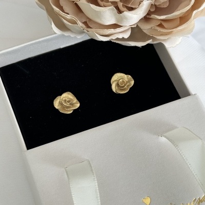 Brincos dourados em forma de flor numa caixa preta e saco branco com texto dourado num fundo branco com flor.