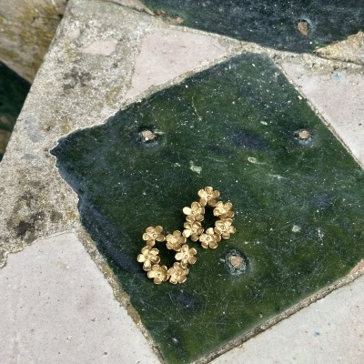 Brincos dourados em forma de flores pequenos sobre mosaico de azulejos verdes e bege.