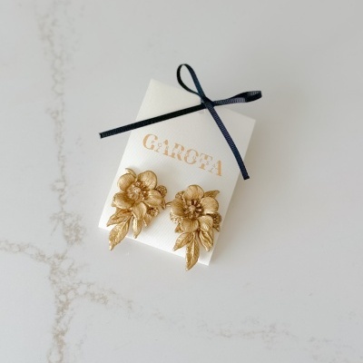 Brincos dourados em forma de flores detalhadas numa embalagem branca com laço preto