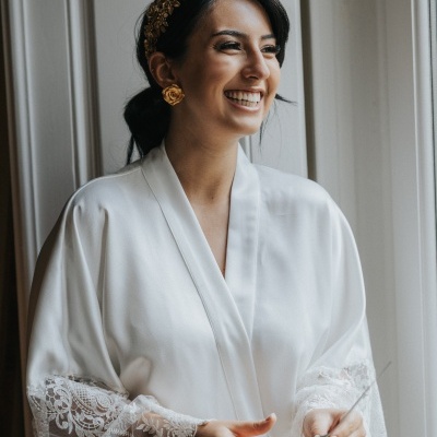 Mulher com roupão branco acetinado e renda, acessórios dourados, sorrindo junto à janela