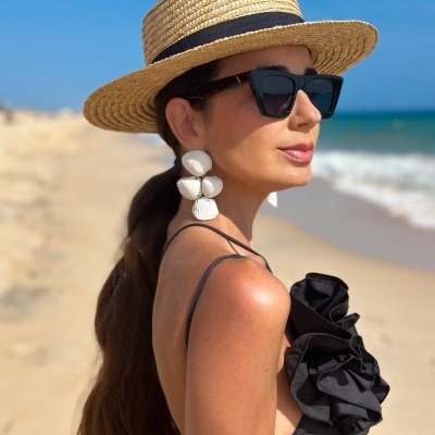 Mulher na praia com chapéu de palha, óculos escuros, brincos de conchas e top preto.