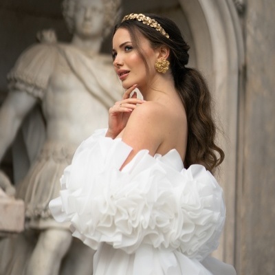 Mulher com vestido branco de mangas volumosas e acessórios dourados, estátua clássica ao fundo