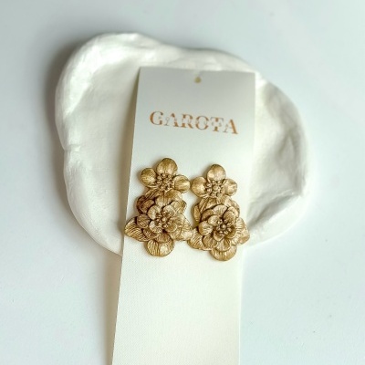 brincos dourados em forma de flores num suporte branco com texto GAROTA