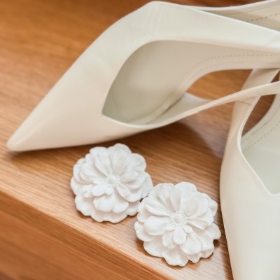 Sapatos de salto alto brancos com bico apontado e acessórios de flores brancas numa superfície de madeira