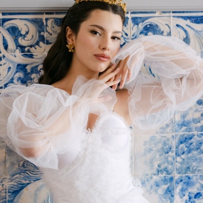 Mulher com vestido de noiva branco de mangas bufantes e acessórios dourados perante azulejos azuis e brancos tradicionais